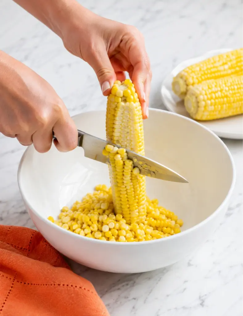 Hands cutting fresh corn kernels off the cob into a white bowl for a corn salad recipe on a marble kitchen countertop.