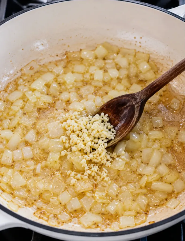 Chopped onions and minced garlic cooking in bacon fat in a white pan for a homemade baked beans recipe.