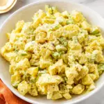 Classic potato salad in a white bowl with creamy dressing, chopped celery, red onion, black pepper, and an orange kitchen towel on a marble countertop.