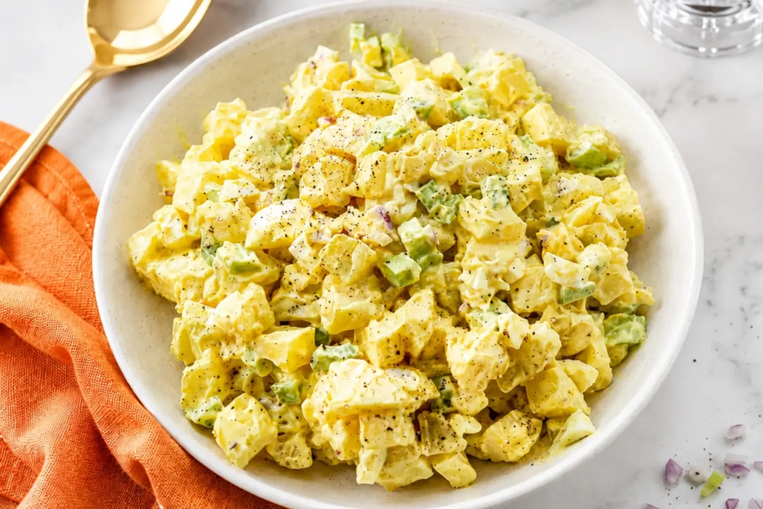 Classic potato salad in a white bowl with creamy dressing, chopped celery, red onion, black pepper, and an orange kitchen towel on a marble countertop.