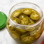 Open jar of Quick Pickled Jalapeños filled with sliced jalapeños in brine, with a green lid and orange towel on a white marble kitchen counter.