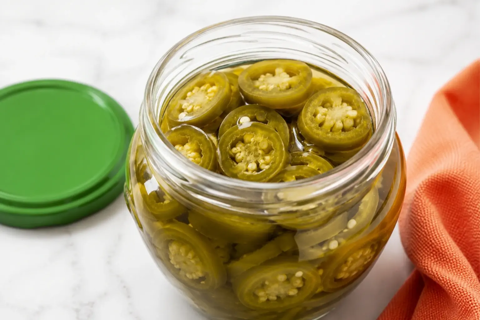 Open jar of Quick Pickled Jalapeños filled with sliced jalapeños in brine, with a green lid and orange towel on a white marble kitchen counter.