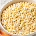 Creamy macaroni salad in a white bowl with celery, red onion, egg, and pepper on a marble kitchen countertop with an orange towel nearby.
