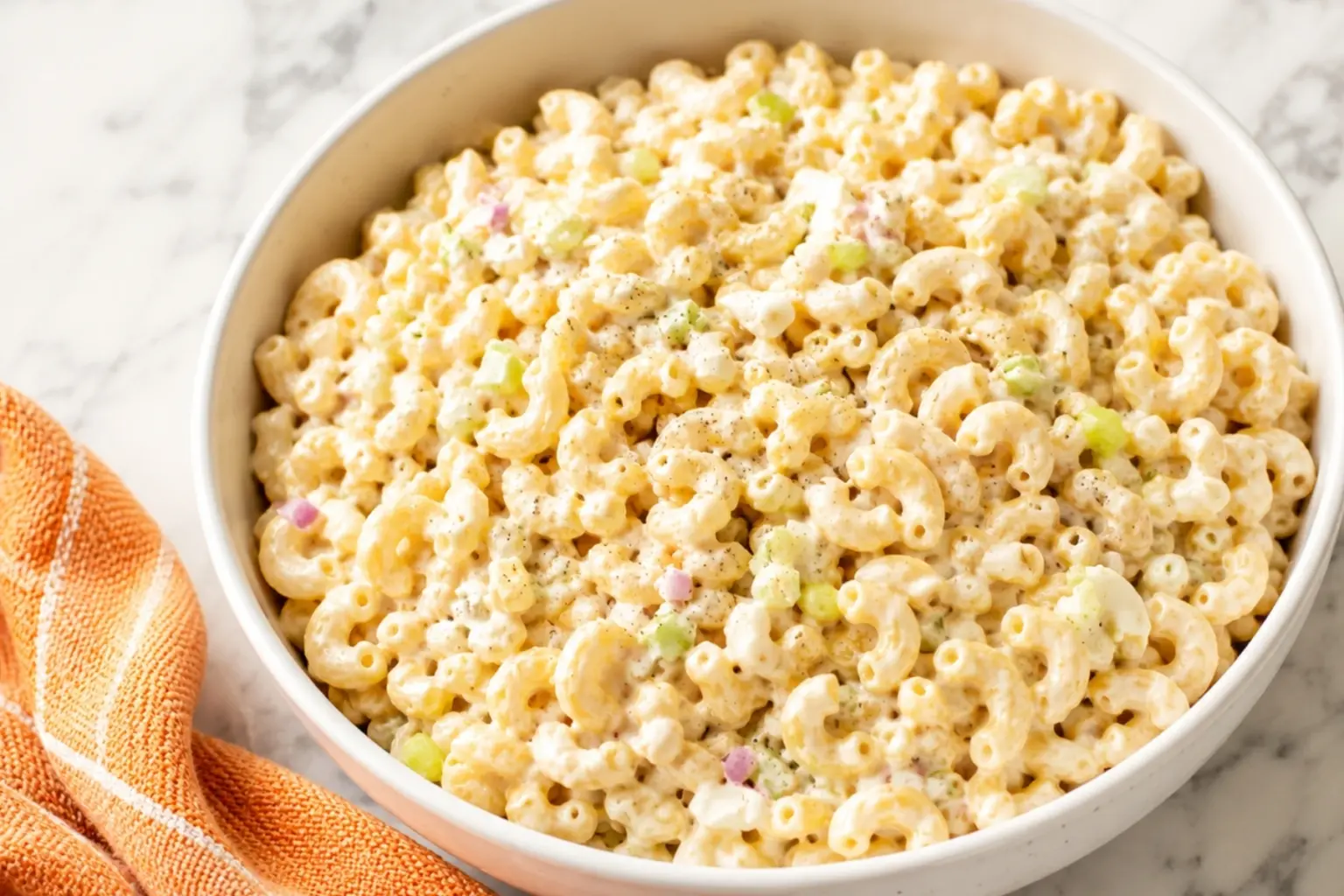 Creamy macaroni salad in a white bowl with celery, red onion, egg, and pepper on a marble kitchen countertop with an orange towel nearby.