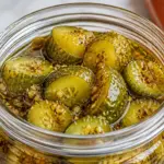 Garlic Dill Pickles in a glass jar with sliced cucumbers, garlic, dill spices, and golden brine on a white marble countertop with an orange kitchen towel.