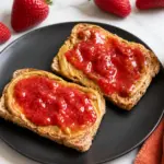 Low Sugar Strawberry Lemon Jam spread over peanut butter toast on a black plate with fresh strawberries and a jam jar on marble countertop.