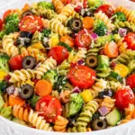 Close-up Pasta Salad with Veggies in a crystal bowl with tri-color rotini, cherry tomatoes, broccoli, black olives, carrots, cucumber, and parmesan on a marble countertop