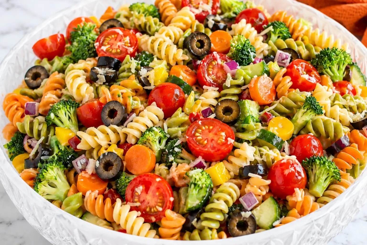 Close-up Pasta Salad with Veggies in a crystal bowl with tri-color rotini, cherry tomatoes, broccoli, black olives, carrots, cucumber, and parmesan on a marble countertop