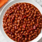 Overhead shot of glossy baked beans in a white bowl on a marble countertop with an orange kitchen towel nearby.