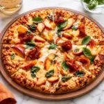 Prosciutto peach pizza with melted cheese, fresh basil, juicy peach slices, and crispy prosciutto on a wooden board over a white marble countertop.