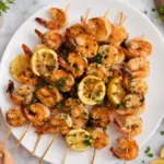 Garlic butter shrimp skewers served on a white plate with grilled lemon slices, fresh parsley, and an orange kitchen towel on a marble countertop.