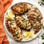 Lemon herb marinated grilled chicken breasts served on a white plate with sliced chicken, lemon wedges, fresh parsley, and an orange kitchen towel on a marble countertop.
