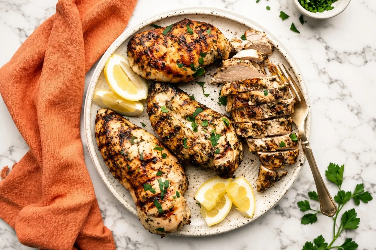 Lemon herb marinated grilled chicken breasts served on a white plate with sliced chicken, lemon wedges, fresh parsley, and an orange kitchen towel on a marble countertop.
