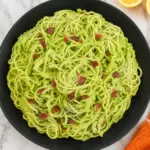 Creamy avocado pasta with crispy bacon in a black bowl on a white marble countertop with lemon and orange towel