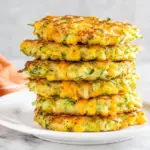 Stack of cheesy zucchini and corn fritters with golden crispy edges on a white plate beside an orange kitchen towel.