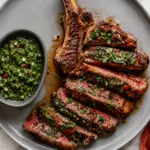 Chimichurri steak sliced medium rare on a gray plate with fresh herb sauce, red chili, garlic, and a side bowl of chimichurri on a white marble countertop.