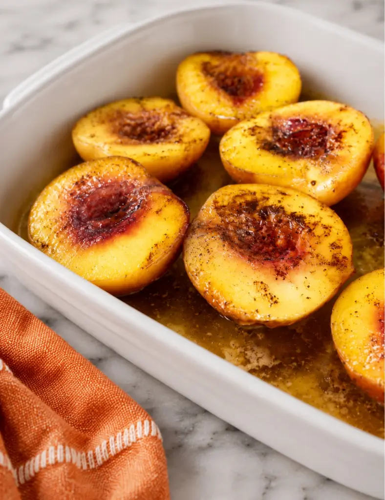 Close-up of baked peaches with honey and cinnamon in a white baking dish with golden syrup and an orange kitchen towel.