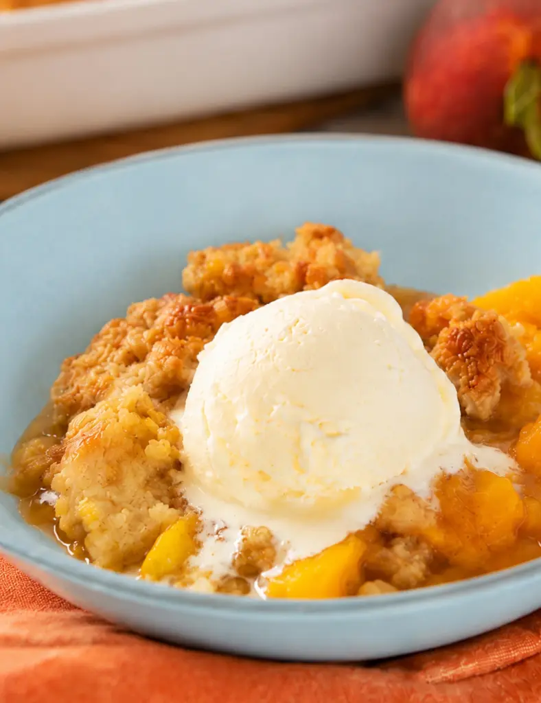 Close-up of Southern Homemade Peach Cobbler with melting vanilla ice cream, juicy peach filling, and golden crumbly topping in a blue bowl.