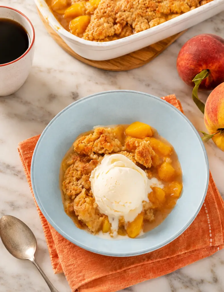 Overhead shot of Southern Homemade Peach Cobbler with vanilla ice cream, juicy peach filling, golden topping, fresh peaches, and orange towel.