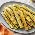 Baked Parmesan Zucchini sticks seasoned with herbs and black pepper on a blue oval plate with an orange kitchen towel