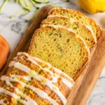 Sliced zucchini bread loaf with lemon glaze on a wooden board, served with fresh lemon and an orange kitchen towel on a marble countertop.