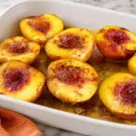 Baked peaches with honey and cinnamon in a white baking dish on a marble countertop with an orange kitchen towel.