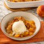 Southern homemade peach cobbler served warm with vanilla ice cream, fresh peaches, and orange kitchen towel on marble countertop.