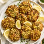 Overhead view of golden Chicken Zucchini Fritters arranged on a white plate with lemon wedges, chopped herbs, and chives on a marble countertop.